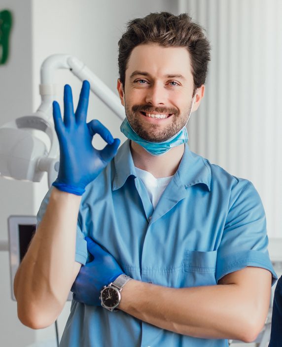 Smiling dentist in blue scrubs making an OK sign
